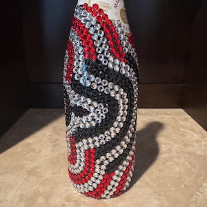 May include: A glass bottle covered in a decorative pattern of small, circular, faceted rhinestones in red, black, and clear colors. The rhinestones are arranged in swirling, wave-like lines that wrap around the body of the bottle.