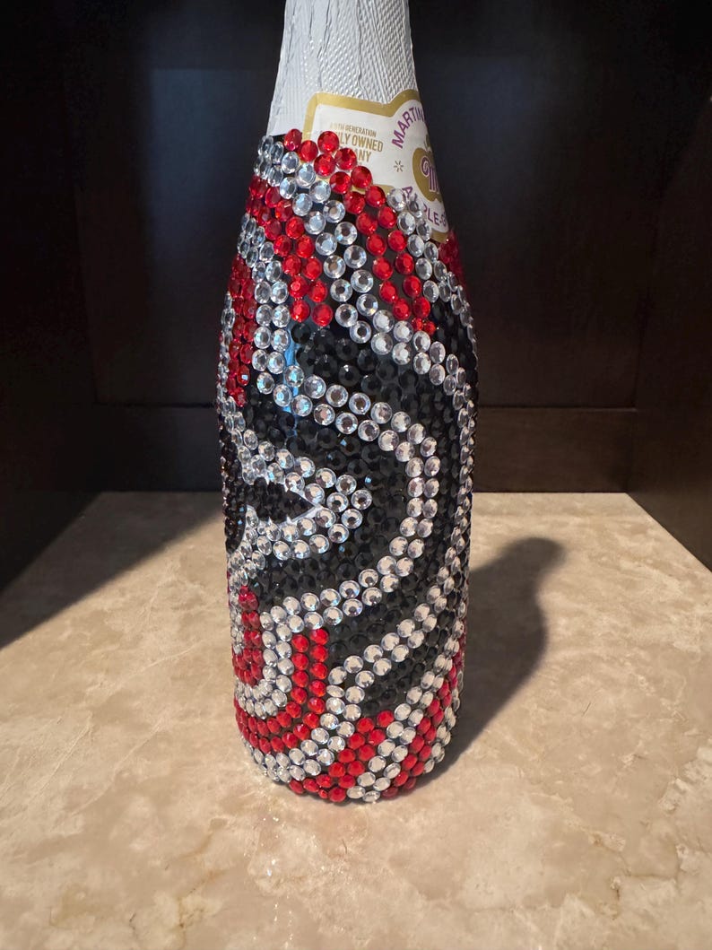 May include: A glass bottle covered in a decorative pattern of small, circular, faceted rhinestones in red, black, and clear colors. The rhinestones are arranged in swirling, wave-like lines that wrap around the body of the bottle.