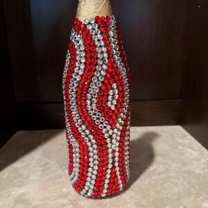 May include: A decorative bottle featuring a gold foil neck and a body completely covered in a repeating wavy pattern of small, sparkling red and clear rhinestones arranged in vertical columns that wrap around the entire cylindrical surface.
