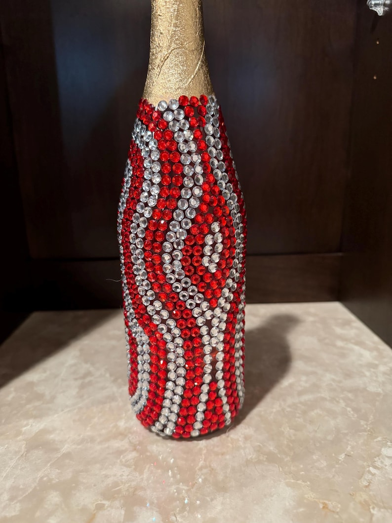 May include: A decorative bottle featuring a gold foil neck and a body completely covered in a repeating wavy pattern of small, sparkling red and clear rhinestones arranged in vertical columns that wrap around the entire cylindrical surface.