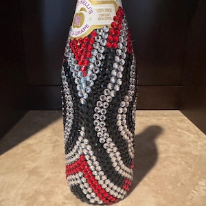 May include: A Martinelli's sparkling grape juice bottle covered in a decorative pattern of red, black, and clear rhinestones. The label at the top remains visible, displaying the text Martinelli's, 100% juice, and contains no alcohol.