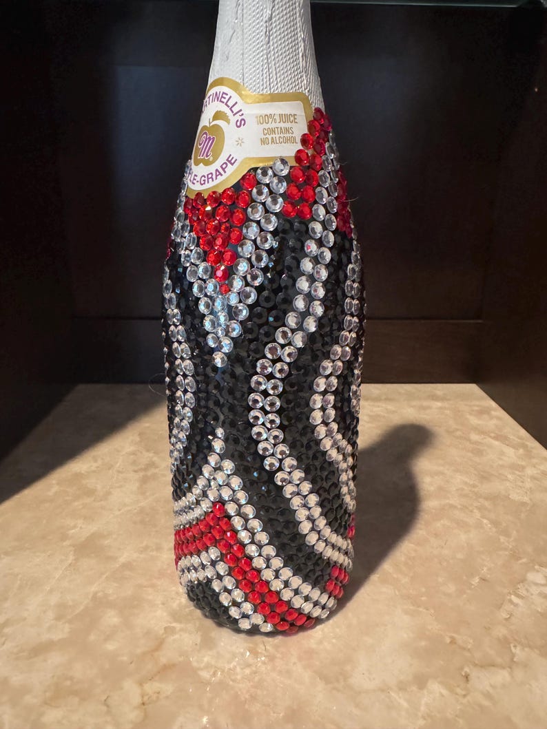 May include: A Martinelli's sparkling grape juice bottle covered in a decorative pattern of red, black, and clear rhinestones. The label at the top remains visible, displaying the text Martinelli's, 100% juice, and contains no alcohol.