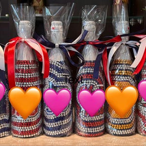 May include: Six glass bottles wrapped in clear plastic, each decorated with rows of colorful rhinestones in various patterns. Each bottle is topped with a white cap and tied with a satin ribbon in red, white, and blue. The word Text is visible.