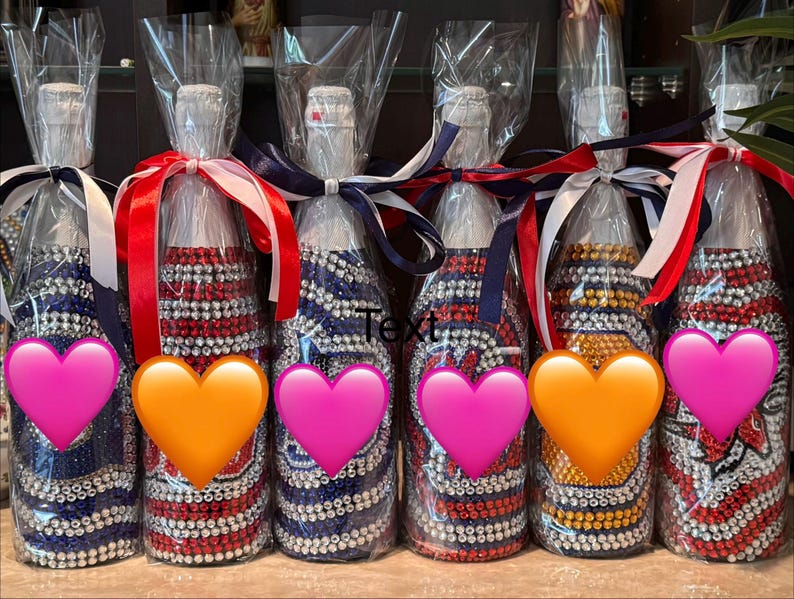 May include: Six glass bottles wrapped in clear plastic, each decorated with rows of colorful rhinestones in various patterns. Each bottle is topped with a white cap and tied with a satin ribbon in red, white, and blue. The word Text is visible.