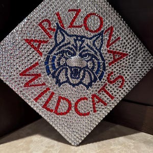 May include: A graduation cap decorated with rhinestones in a blue, red, and silver pattern. The cap features the words "Arizona Wildcats" and a stylized image of a wildcat.