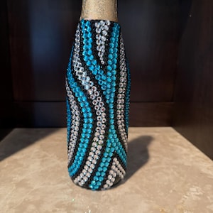 May include: A glass bottle covered in a decorative pattern of small, reflective rhinestones in shades of bright turquoise, clear white, and black. The neck of the bottle is wrapped in a metallic gold foil finish, contrasting with the jeweled body.