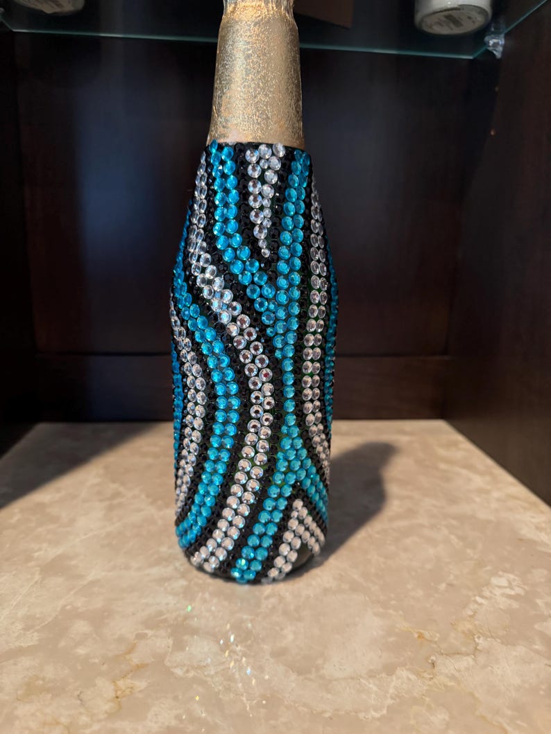 May include: A glass bottle covered in a decorative pattern of small, reflective rhinestones in shades of bright turquoise, clear white, and black. The neck of the bottle is wrapped in a metallic gold foil finish, contrasting with the jeweled body.