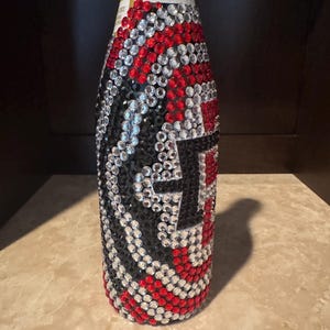 May include: A bottle completely covered in a decorative pattern of small, round, reflective rhinestones in alternating colors of red, black, and clear. The stones are arranged in a swirling, wave-like design that wraps around the entire exterior.