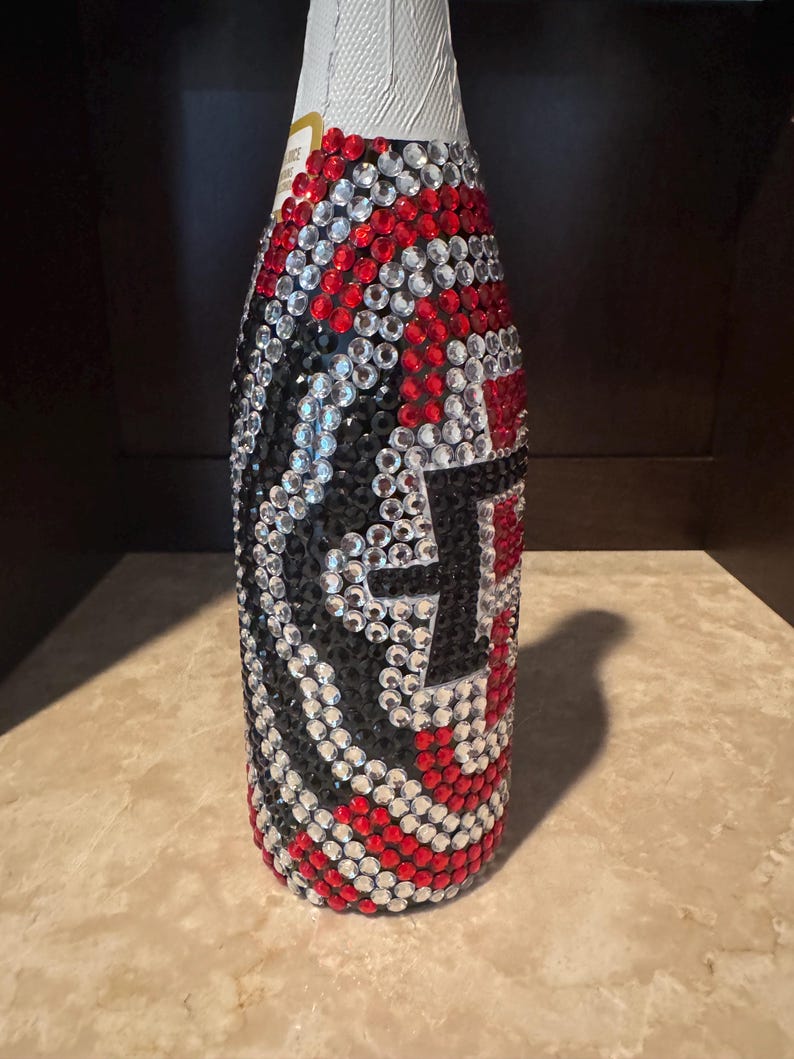 May include: A bottle completely covered in a decorative pattern of small, round, reflective rhinestones in alternating colors of red, black, and clear. The stones are arranged in a swirling, wave-like design that wraps around the entire exterior.
