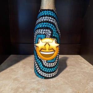 May include: A glass bottle covered in a swirling pattern of blue and clear rhinestones on a black base. The neck of the bottle is wrapped in gold foil. A star-eyed smiling emoji is positioned over the center of the bottle, obscuring the design.