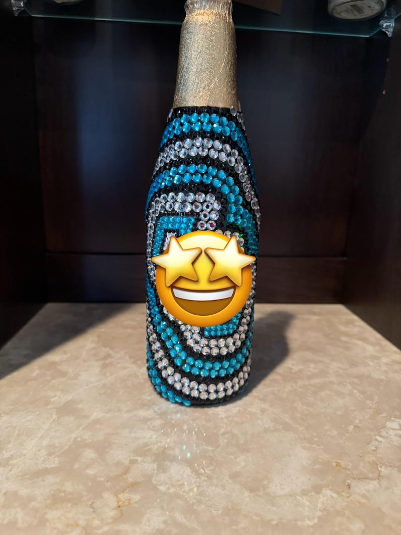 May include: A glass bottle covered in a swirling pattern of blue and clear rhinestones on a black base. The neck of the bottle is wrapped in gold foil. A star-eyed smiling emoji is positioned over the center of the bottle, obscuring the design.