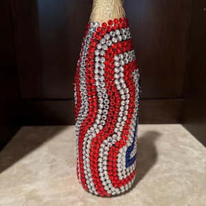 May include: A glass bottle with a gold foil neck, fully covered in a decorative pattern of small, sparkling red, white, and blue rhinestones arranged in vertical wavy lines that wrap around the entire cylindrical body of the container.