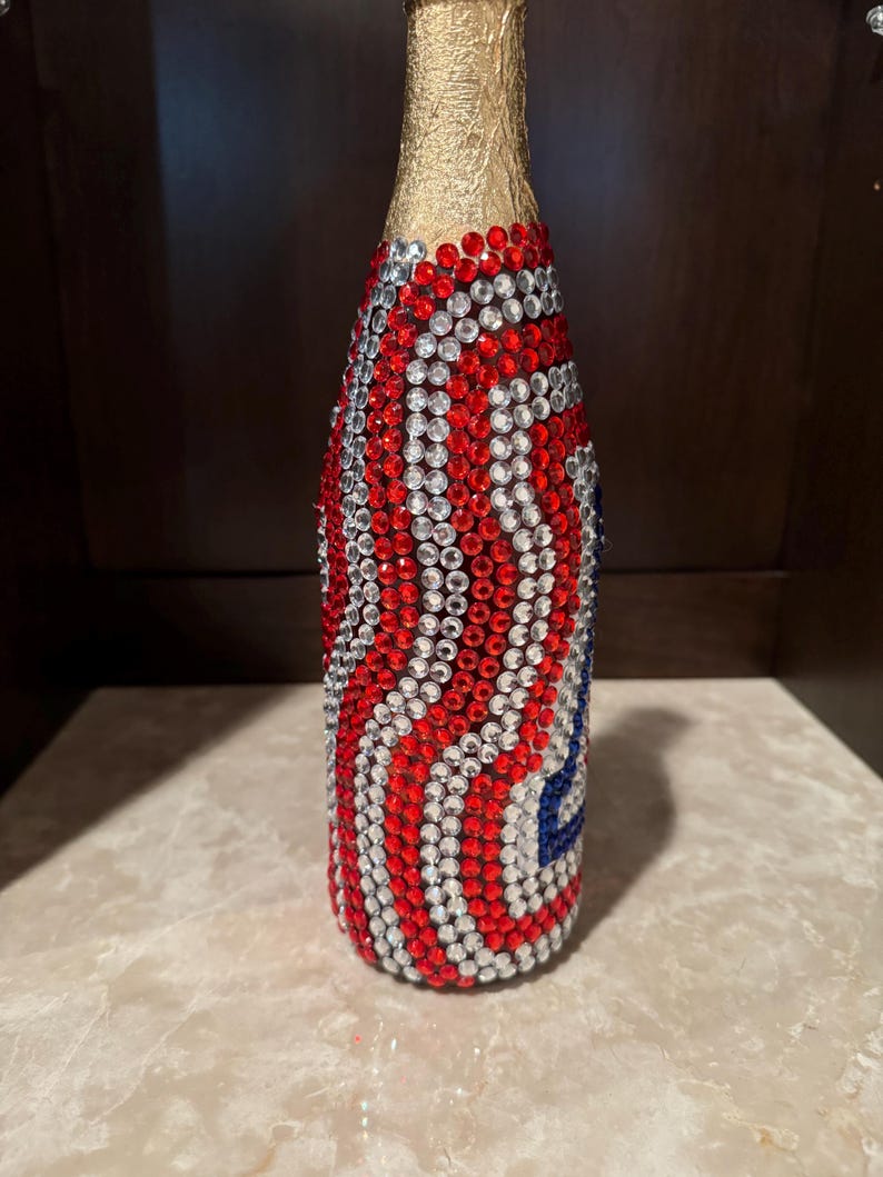May include: A glass bottle with a gold foil neck, fully covered in a decorative pattern of small, sparkling red, white, and blue rhinestones arranged in vertical wavy lines that wrap around the entire cylindrical body of the container.