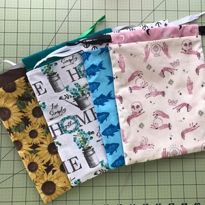 May include: A collection of fabric pouches with various patterns. Designs include sunflowers, a farmhouse theme with text, blue fish, and a pink and white pattern with skulls, hands, and crystals. Each pouch has a drawstring.