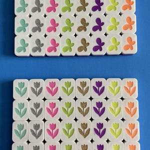 May include: Two sets of white tiles with colorful butterfly and tulip designs. The butterflies are in shades of teal, silver, pink, green, gold, purple, and orange. The tulips are teal, silver, pink, green, gold, purple, and orange. The background is a solid blue.