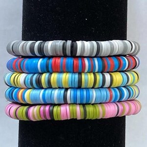 May include: Stack of six colorful bracelets. Each bracelet is made of small, flat, round beads. The bracelets feature a variety of colors, including gray, black, red, blue, yellow, and pink. The bracelets are stacked on a black surface.