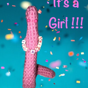 May include: A pink crocheted figure with pink eyes and a necklace spelling "It's a Girl" against a teal background with confetti. The text "It's a Girl!!!" is written in pink. A whimsical announcement for a baby girl.