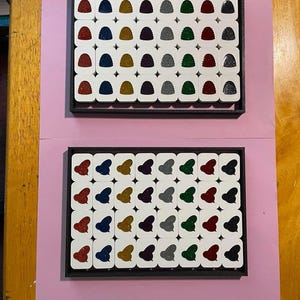 May include: Two rectangular black trays containing rows of small white tiles. The top tray features tiles with glittery, rounded shapes in red, blue, gold, purple, silver, and green. The bottom tray holds tiles with glittery heart-shaped cutouts.