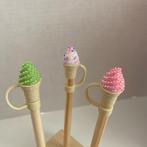 Op de afbeelding: Drie decoratieve potloden met ijsjestoppers. De ijsjes zijn groen, roze en wit, elk versierd met sprankelende versieringen. De potloden worden in een houten standaard gehouden.