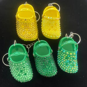 Puede incluir: Cinco llaveros en miniatura con forma de zapato. Dos son amarillos con pedrería transparente y tres son verdes con pedrería iridiscente.