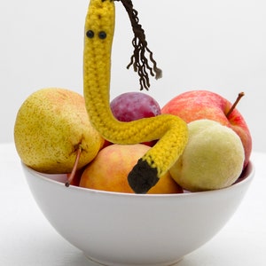 May include: A white ceramic bowl filled with various fruits, including a pear, apple, peach, and plum. A yellow crocheted banana-shaped figure with a brown top and eyes is placed among the fruit.