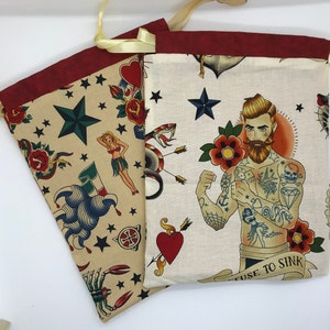 May include: Two fabric drawstring bags with a red trim. The bags are decorated with a variety of colorful tattoo-style designs, including stars, hearts, snakes, anchors, and a man with tattoos. One bag has the text "Refuse to Sink".