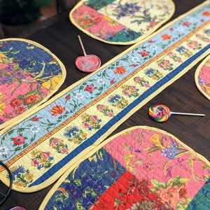 K&ouml;nnte beinhalten: Ein Set aus bunten, gesteppten Platzsets mit Blumenmuster und einem passenden Tischl&auml;ufer. Die Platzsets und der Tischl&auml;ufer haben ein Patchwork-Design mit rosa, blauen, gelben und gr&uuml;nen Blumenmustern. Die Platzsets sind oval geformt und der Tischl&auml;ufer ist rechteckig.
