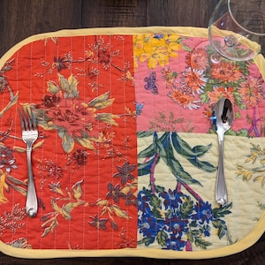 Handcrafted patchwork placemat featuring a vibrant mix of floral designs in red, pink, yellow, and green sections. The placemat is bordered with a yellow trim, showcasing its quality and of charm to any dining table. Made in the USA  Bohemian style patchwork placemats with floral motifs and yellow trim, styled with white china and silverware for elegant table settings