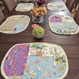 May include: A wooden table set with six place settings. Each place setting includes a quilted placemat with a floral pattern, a fork, and a spoon. The table is dark brown and has a distressed finish.