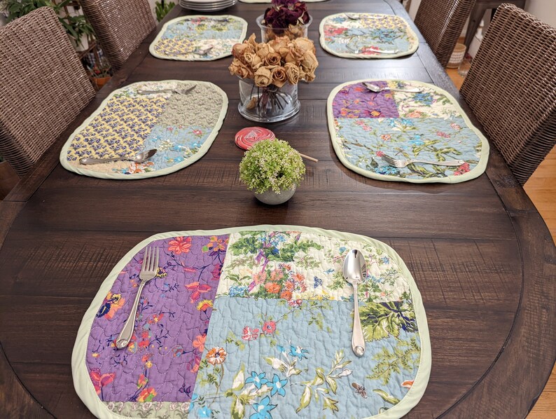 May include: A wooden table set with six place settings. Each place setting includes a quilted placemat with a floral pattern, a fork, and a spoon. The table is dark brown and has a distressed finish.