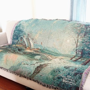 May include: A white couch with a blue and green tapestry throw blanket featuring a winter scene with a bridge, trees, and a house. The blanket has a fringe border and the signature "Thomas Kinkade" is visible in the bottom right corner.