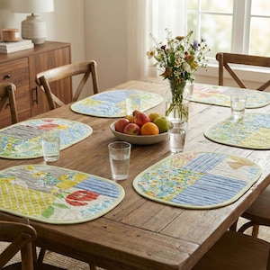 May include: A rustic wooden dining table set with six oval placemats. Each placemat features a patchwork design with floral and geometric patterns in shades of blue, yellow, and red. A bowl of fruit and a vase of flowers are in the center.