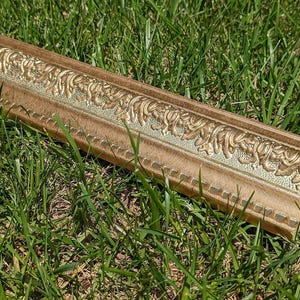May include: A section of ornate, gold-colored picture frame molding with a detailed, raised floral design. The frame has a light brown base and is resting on green grass.