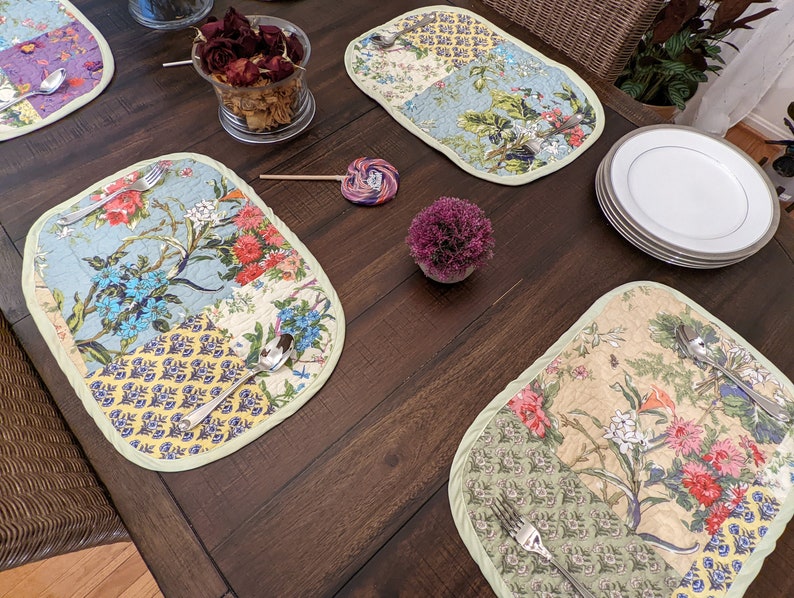 May include: Four quilted placemats with floral patterns in shades of blue, green, yellow, and pink. The placemats are on a wooden table with a vase of dried flowers, a lollipop, and a small potted plant.