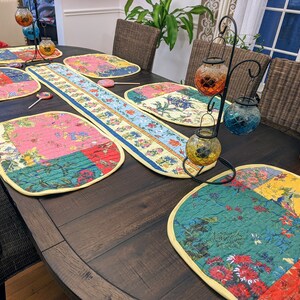 K&ouml;nnte beinhalten: Ein dunkler Holztisch, der mit einem gelben und wei&szlig;en Tischl&auml;ufer und bunten gesteppten Platzsets gedeckt ist. Der Tischl&auml;ufer hat ein Blumenmuster und die Platzsets haben ein Patchwork-Design mit Blumen- und geometrischen Mustern. Es gibt auch mehrere dekorative Kerzenhalter mit Glaskugeln.