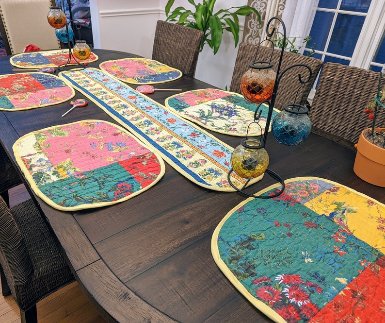 K&ouml;nnte beinhalten: Ein dunkler Holztisch, der mit einem gelben und wei&szlig;en Tischl&auml;ufer und bunten gesteppten Platzsets gedeckt ist. Der Tischl&auml;ufer hat ein Blumenmuster und die Platzsets haben ein Patchwork-Design mit Blumen- und geometrischen Mustern. Es gibt auch mehrere dekorative Kerzenhalter mit Glaskugeln.