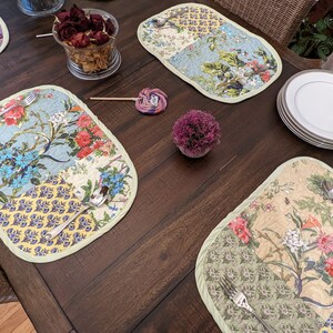 Four quilted placemats in floral patterns, featuring blue, green, yellow, and pink hues.  Each mat has a light green border. Silverware rests on two of the placemats.  White plates are stacked on the table.  The placemats are rectangular with rounded corners, showcasing a patchwork design with various floral motifs.  The overall aesthetic is charming and suitable for a dining setting.