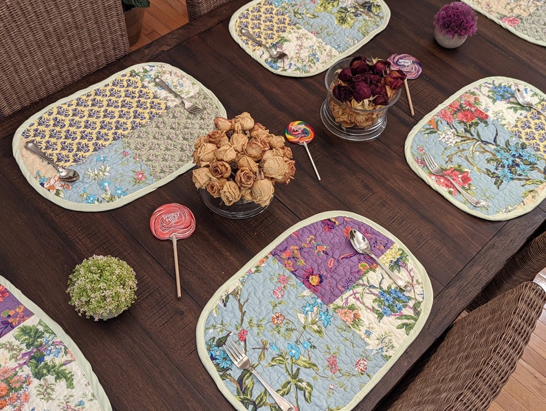 May include: A dark wood table set with four oval placemats featuring a patchwork design of floral patterns in shades of blue, green, yellow, and purple. The placemats are accented with a light green border. A bowl of dried roses, two lollipops, and a small potted plant are also on the table.