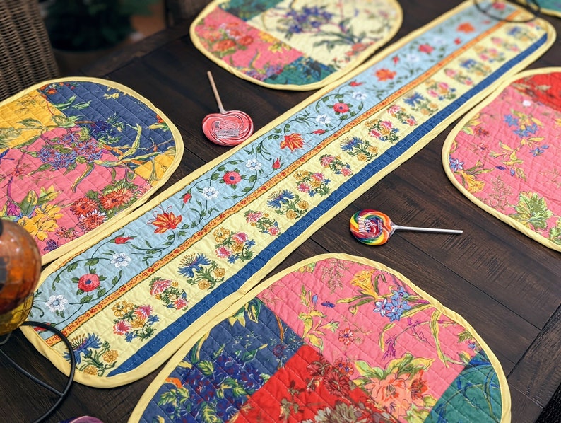 K&ouml;nnte beinhalten: Ein Set aus bunten, gesteppten Platzsets mit Blumenmuster und einem passenden Tischl&auml;ufer. Die Platzsets und der Tischl&auml;ufer haben ein Patchwork-Design mit rosa, blauen, gelben und gr&uuml;nen Blumenmustern. Die Platzsets sind oval geformt und der Tischl&auml;ufer ist rechteckig.
