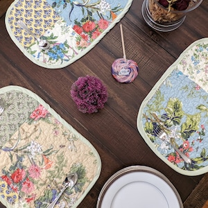 May include: A table setting with four placemats featuring a floral patchwork design. Each placemat has a green border and features a variety of flowers and leaves in shades of blue, yellow, green, and pink. The placemats are on a dark brown wooden table.