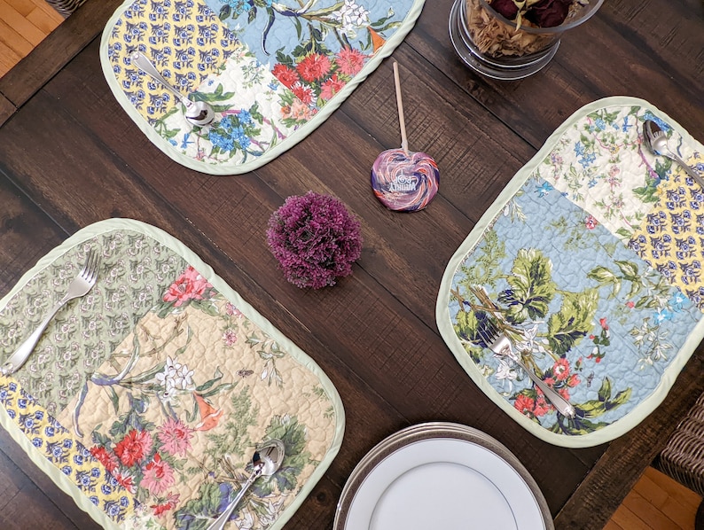 May include: A table setting with four placemats featuring a floral patchwork design. Each placemat has a green border and features a variety of flowers and leaves in shades of blue, yellow, green, and pink. The placemats are on a dark brown wooden table.