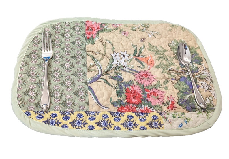 May include: A green quilted placemat with a floral pattern featuring pink, red, yellow, and blue flowers. A silver fork and spoon are placed on the placemat.