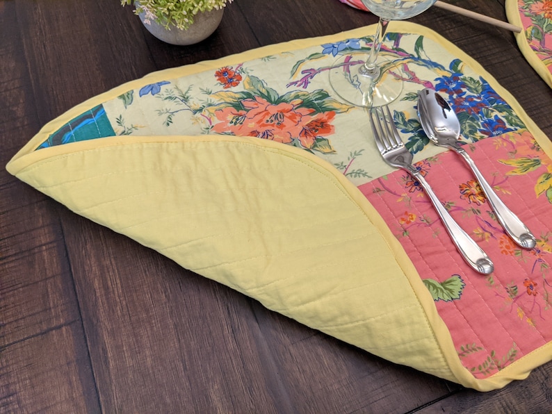 Vibrant handcrafted placemat featuring a patchwork of floral designs in red, pink, yellow, and green, bordered with a cheerful yellow trim. This elegant piece is ideal for brightening up any dining experience. Proudly made in the USA. Eclectic patchwork placemats set combining vintage floral prints and solid color blocks, finished with sunny yellow binding for lively table décor