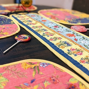 May include: A set of four quilted placemats and a table runner with a floral pattern in shades of pink, yellow, blue, and green. The placemats and runner have a yellow border.