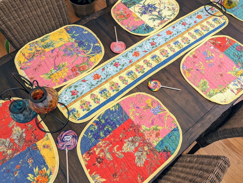 K&ouml;nnte beinhalten: Ein Set aus bunten, gesteppten Platzsets mit Blumenmuster und einem passenden Tischl&auml;ufer. Die Platzsets sind oval geformt und der L&auml;ufer ist rechteckig. Das Blumenmuster ist eine Mischung aus Rosa, Gelb, Blau und Gr&uuml;n.