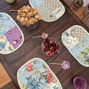 May include: A dark wood table setting with four oval placemats featuring a patchwork design of blue, green, purple, and yellow floral patterns. The placemats have a light green border. There are two sets of silverware on the placemats, and a bowl of dried rose petals in the center of the table.