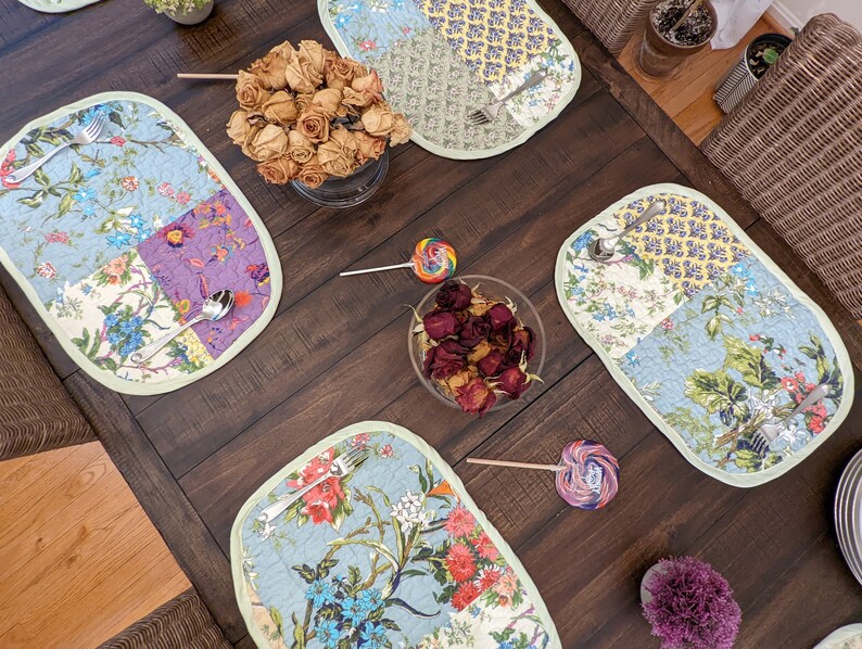May include: A dark wood table setting with four oval placemats featuring a patchwork design of blue, green, purple, and yellow floral patterns. The placemats have a light green border. There are two sets of silverware on the placemats, and a bowl of dried rose petals in the center of the table.