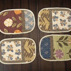 May include: Four oval quilted placemats with various patterns. Each mat features a different design, including floral and leaf motifs, in shades of brown, green, and cream. The placemats have a light beige border and are arranged on a dark wood surface.