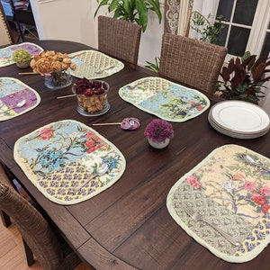 Six quilted placemats in a floral pattern, featuring various shades of green, blue, and pink flowers.  The placemats are arranged on a dark brown wooden table, along with plates, silverware, and small floral arrangements. The placemats have a patchwork design with a light green border.  They are rectangular and appear to be made of a soft fabric.