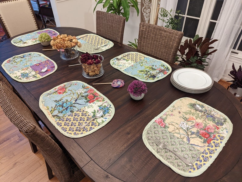 Six quilted placemats in a floral pattern, featuring various shades of green, blue, and pink flowers.  The placemats are arranged on a dark brown wooden table, along with plates, silverware, and small floral arrangements. The placemats have a patchwork design with a light green border.  They are rectangular and appear to be made of a soft fabric.